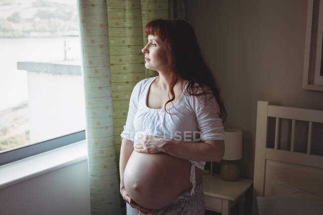 Thoughtful pregnant woman looking through window in bedroom at home — Stock Photo