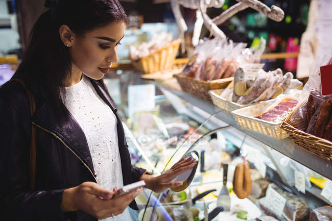 Mujer mirando la exhibición de carne mientras usa el teléfono móvil en el supermercado - foto de stock