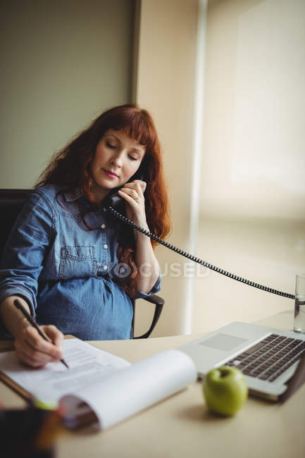 Беременная деловая женщина разговаривает по телефону во время работы в офисе — стоковое фото