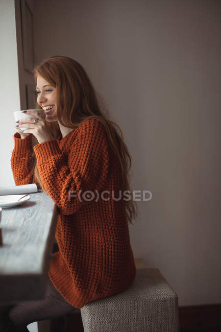 Pelirroja sonriendo mientras toma café en la ventana del restaurante - foto de stock