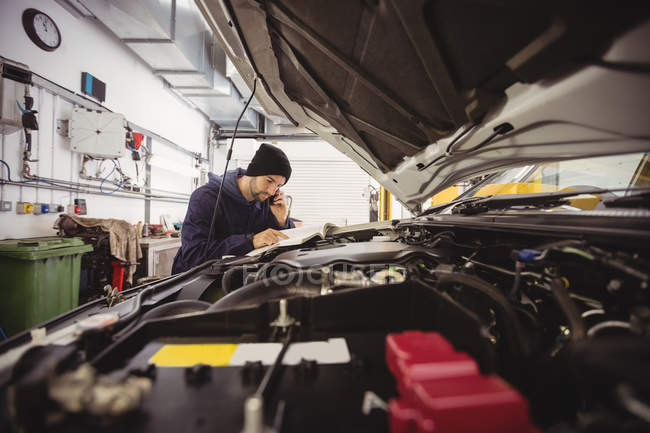 Mechanic reading instruction manual while talking on mobile phone at repair garage — Stock Photo