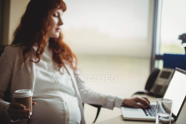 Donna d'affari incinta che utilizza il computer portatile mentre prende il caffè in ufficio — Foto stock