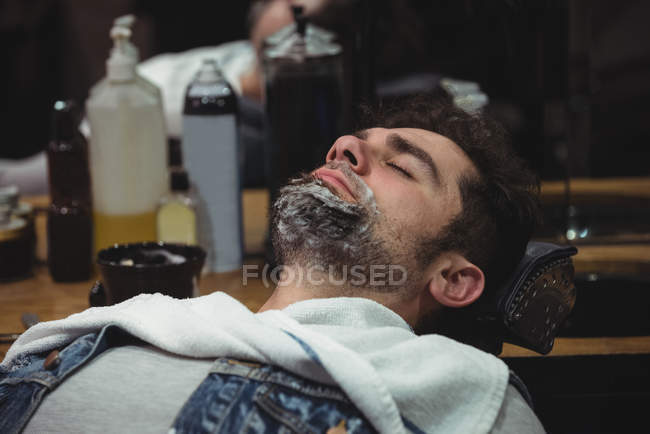 Homme avec crème à raser sur la barbe relaxant sur la chaise dans le salon de coiffure — Photo de stock