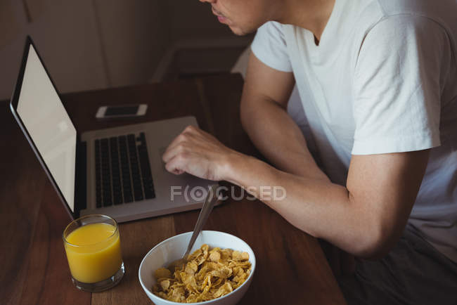 Hombre usando el teléfono móvil y el ordenador portátil mientras desayuna en el dormitorio en casa - foto de stock