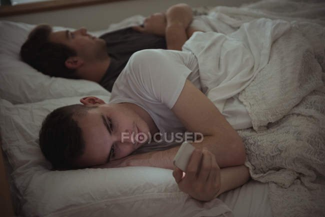 Hombre revisando el teléfono móvil mientras está acostado en la cama con su pareja gay en el dormitorio - foto de stock
