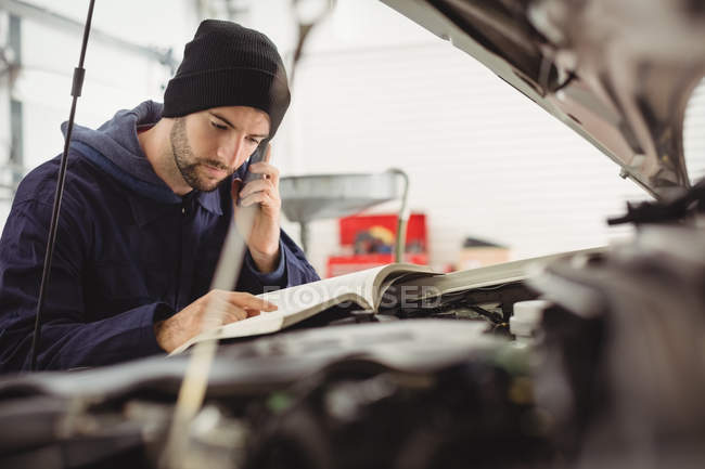 Mechanic reading instruction manual while talking on mobile phone at repair garage — Stock Photo