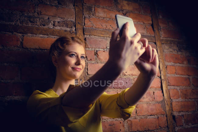 Mujer de pie contra la pared de ladrillo y tomando una selfie en su teléfono móvil - foto de stock