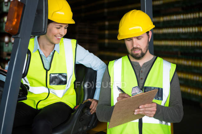 Trabajadora sonriente mirando al trabajador masculino escribiendo en el portapapeles en el almacén - foto de stock