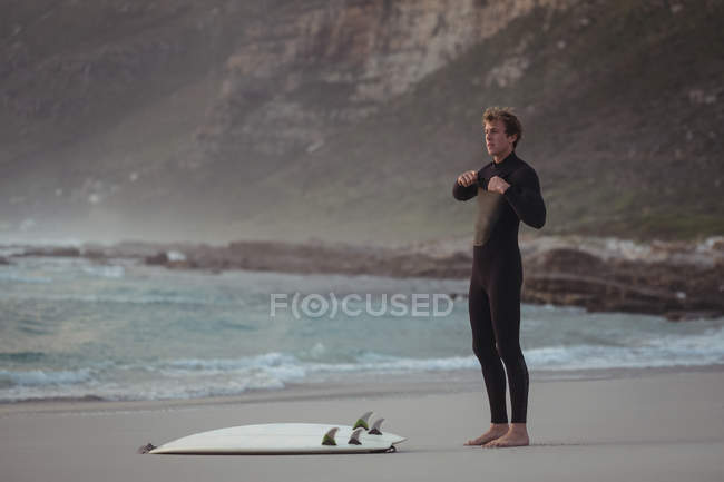 Мужчина в гидрокостюме стоит на пляже с доской для серфинга — стоковое фото