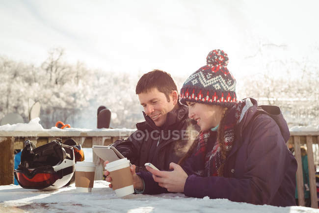 Щаслива пара лижників сидить за столом, використовуючи мобільний телефон та цифровий планшет у гірськолижному курорті — стокове фото