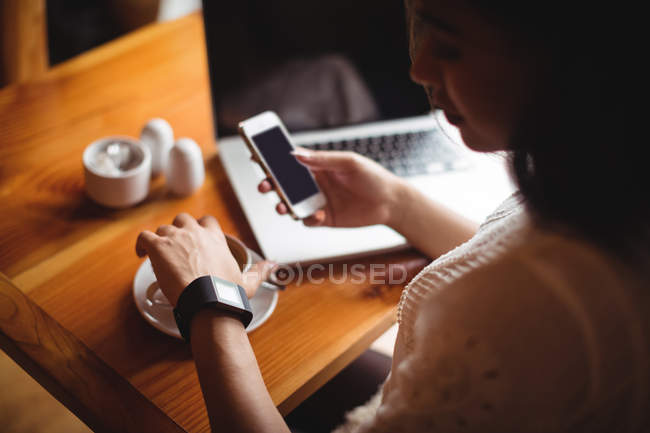 Mujer usando el teléfono móvil mientras toma una taza de café en la cafetería - foto de stock