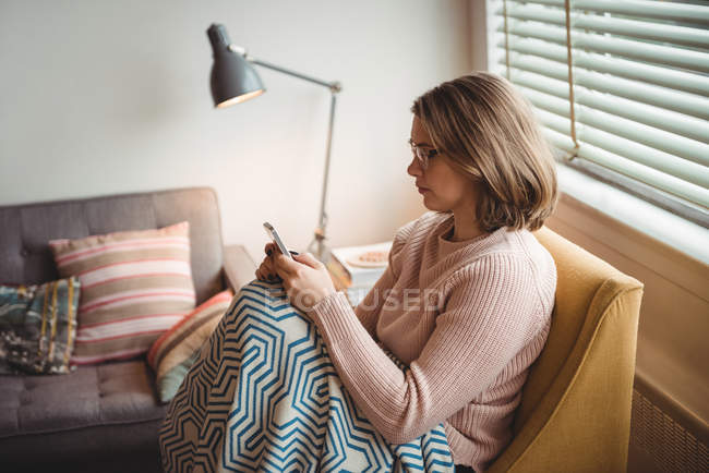 Mulher sentada na cadeira usando telefone celular em casa — Fotografia de Stock