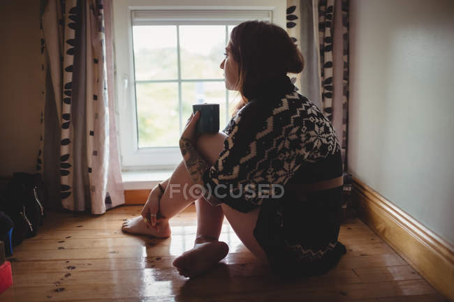 Femme réfléchie assise près de la fenêtre et prenant un café à la maison — Photo de stock