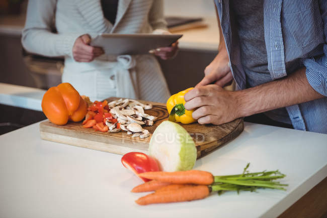 Couple utilisant une tablette numérique tout en hachant des légumes dans la cuisine à la maison — Photo de stock