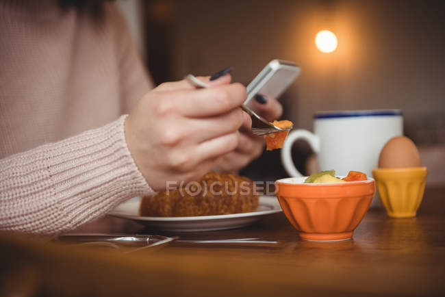 Woman using mobile phone while having breakfast in living room at home — Stock Photo
