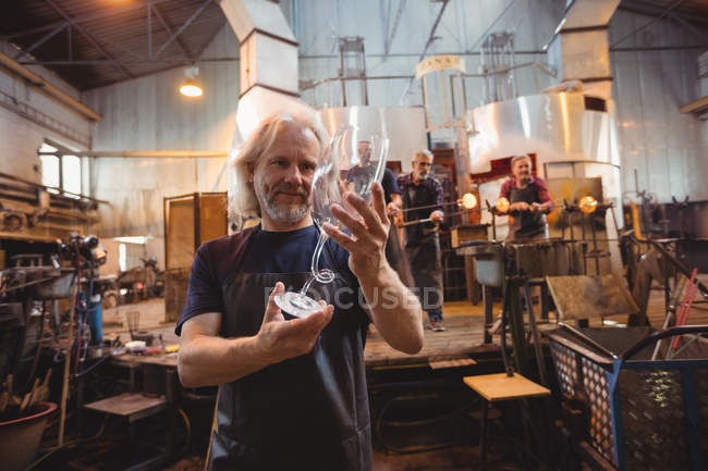 Скляний блендер вивчає скляний посуд, коли колеги працюють у фоновому режимі на скляній фабриці — стокове фото
