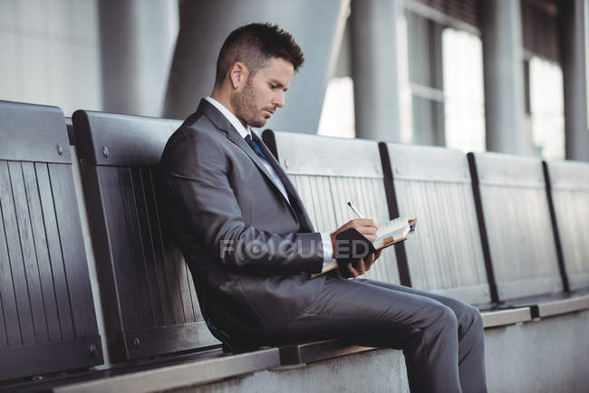 Homme d'affaires écrivant dans un journal alors qu'il était assis sur un banc sur le campus du bureau — Photo de stock
