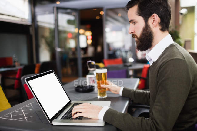 Людина використовує ноутбук, маючи келих пива в барі — стокове фото