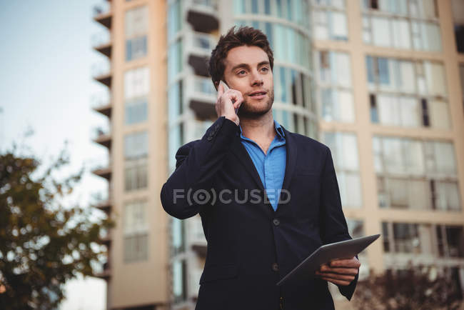 Businessman talking on mobile phone and holding digital tablet near office building — Stock Photo