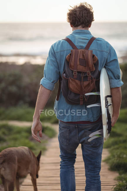 Rear view of a man carrying a surfboard walking on boardwalk — Stock Photo