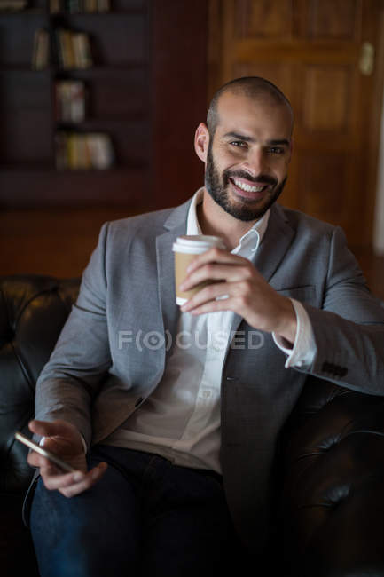 Retrato del hombre de negocios sonriente sosteniendo teléfono móvil y taza de café en la zona de espera en la terminal del aeropuerto - foto de stock