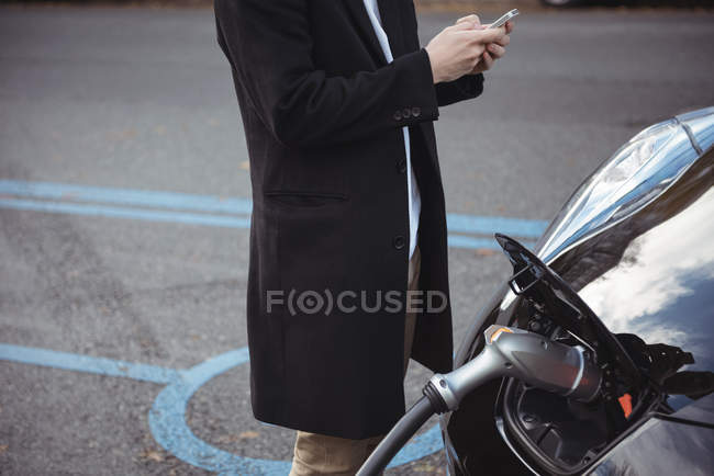 Seção média do homem que usa o telefone móvel ao carregar o carro na estação de carregamento do veículo elétrico — Fotografia de Stock