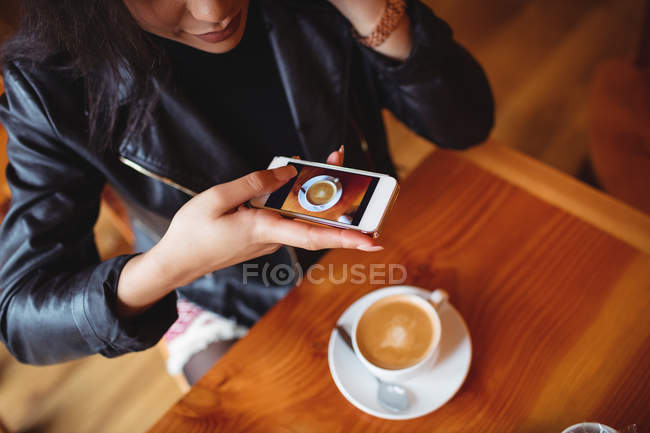 Mujer haciendo clic en la foto de café desde el teléfono móvil en la cafetería - foto de stock