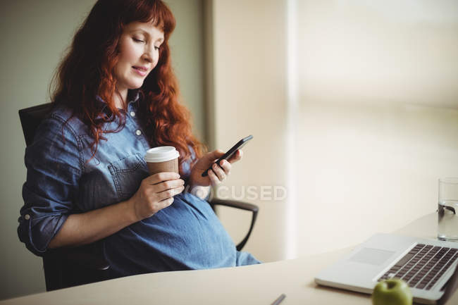 Вагітна бізнес-леді використовує мобільний телефон, маючи каву в офісі — стокове фото