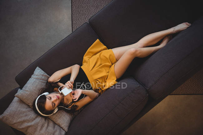 Frau liegt auf Sofa, während sie zu Hause mit Handy Musik hört — Stockfoto