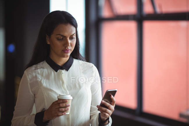 Femme d'affaires tenant tasse de café jetable et en utilisant le téléphone mobile dans le bureau — Photo de stock