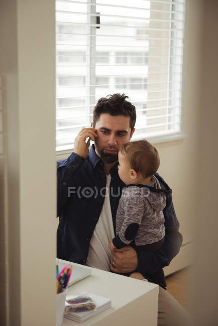 Padre che parla al cellulare mentre tiene in braccio il bambino a casa — Foto stock