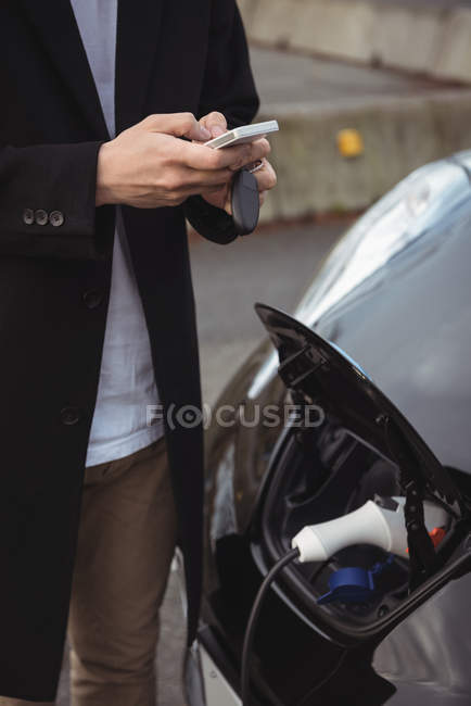Sección media del hombre usando el teléfono móvil mientras carga el coche eléctrico en la calle - foto de stock