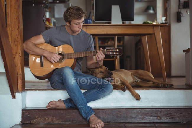 Мужчина играет на гитаре дома, собака лежит рядом с ним — стоковое фото