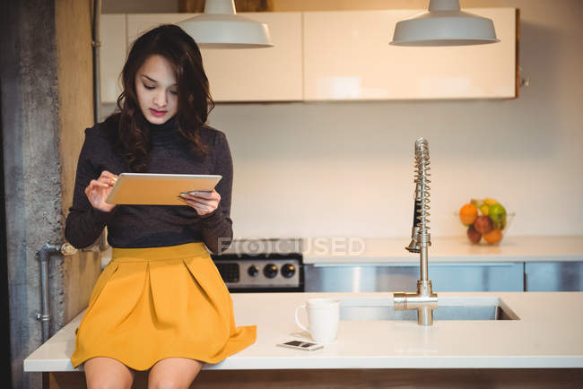 Mujer sentada en la encimera de la cocina usando tableta digital en casa - foto de stock