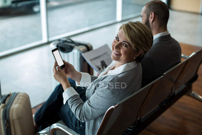 Portrait of smiling businesswoman with mobile phone sitting in waiting area at airport terminal — Stock Photo
