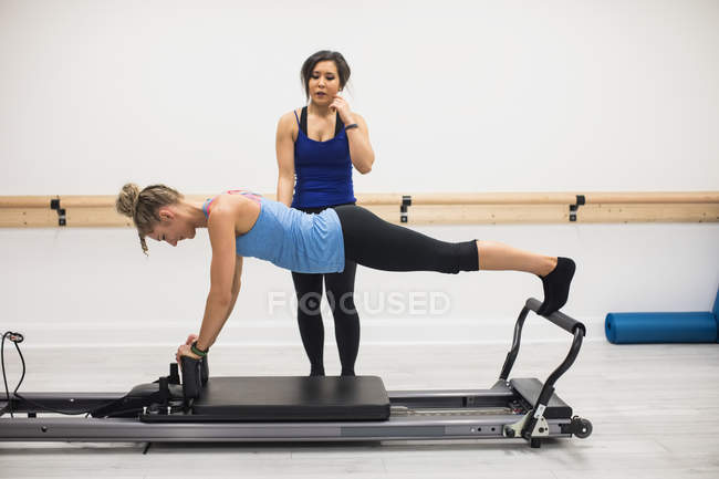 Entrenadora mujer que ayuda a la mujer con el ejercicio de estiramiento en reformador en el gimnasio - foto de stock
