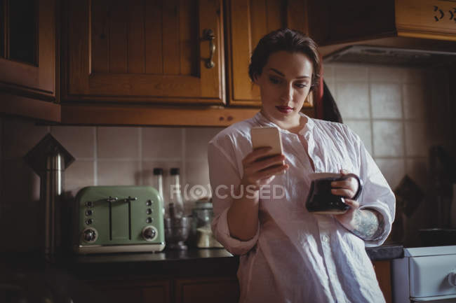 Mulher bonita usando telefone celular enquanto toma café na cozinha em casa — Fotografia de Stock
