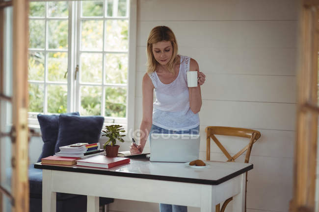 Bella donna che scrive nel diario mentre prende un caffè in soggiorno a casa — Foto stock