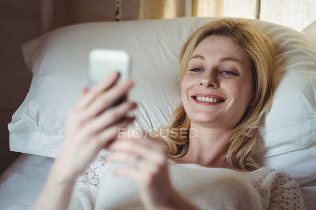 Mulher bonita deitada na cama e usando telefone celular no quarto em casa — Fotografia de Stock