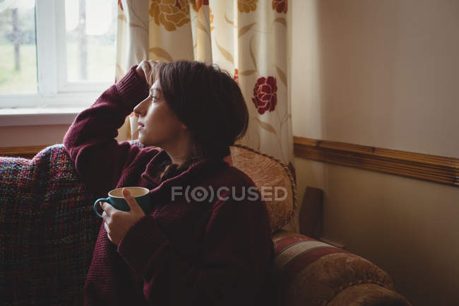 Mujer pensativa sentada y sosteniendo una taza de café en casa - foto de stock