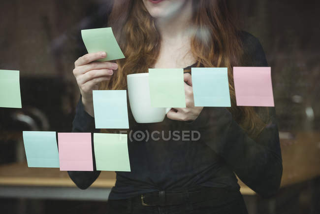 Business executive looking at sticky note while having cup of coffee in office — Stock Photo