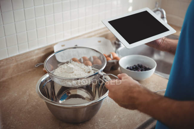 Homem peneirando farinha em uma tigela de mistura enquanto usa um tablet digital na cozinha em casa — Fotografia de Stock