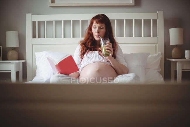 Mulher grávida bebendo suco enquanto lê livro na cama no quarto — Fotografia de Stock