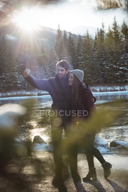 Пара, що стоїть на річці і приймає селфі, використовуючи мобільний телефон взимку — стокове фото