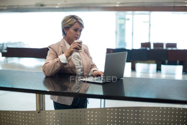 Empresária usando laptop enquanto toma café na área de espera no terminal do aeroporto — Fotografia de Stock