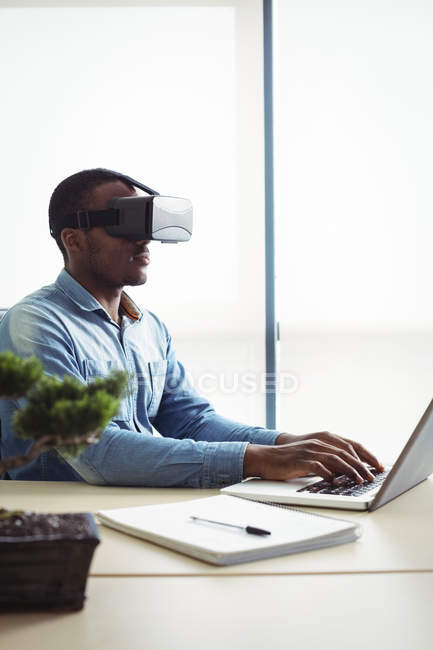 Business executive using virtual reality headset and working on laptop in office — Stock Photo