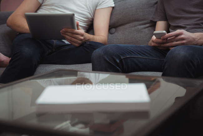 Partie médiane de deux hommes utilisant une tablette numérique et un téléphone portable dans le salon à la maison — Photo de stock