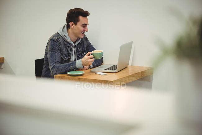 Чоловік дивиться на ноутбук, тримаючи чашку кави в кав'ярні — стокове фото