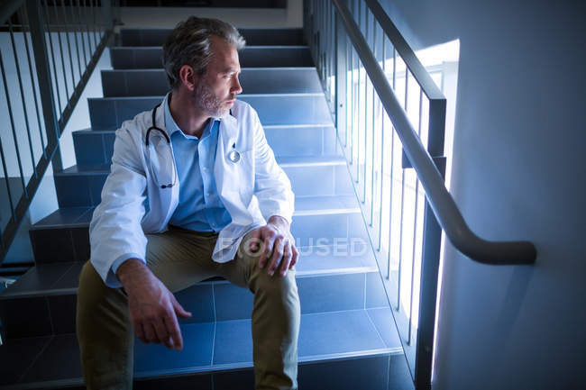 Médico reflexivo sentado en la escalera en el hospital - foto de stock