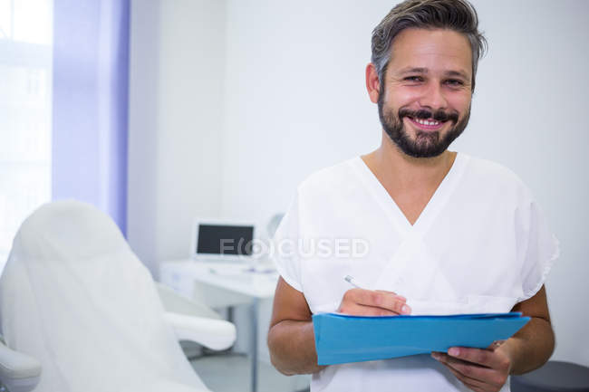 Retrato del médico sonriente escribiendo en informe médico en clínica - foto de stock